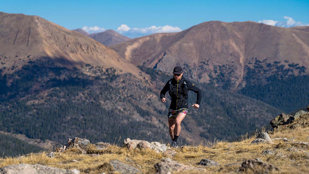 A Male Trail Runner Cresting A Ridge With A Series Of Mountains In The Background Under Blue Skies